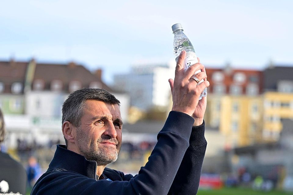 Michael Köllner klatscht im Grünwalder Stadion und hält eine Flasche Wasser in der Hand.