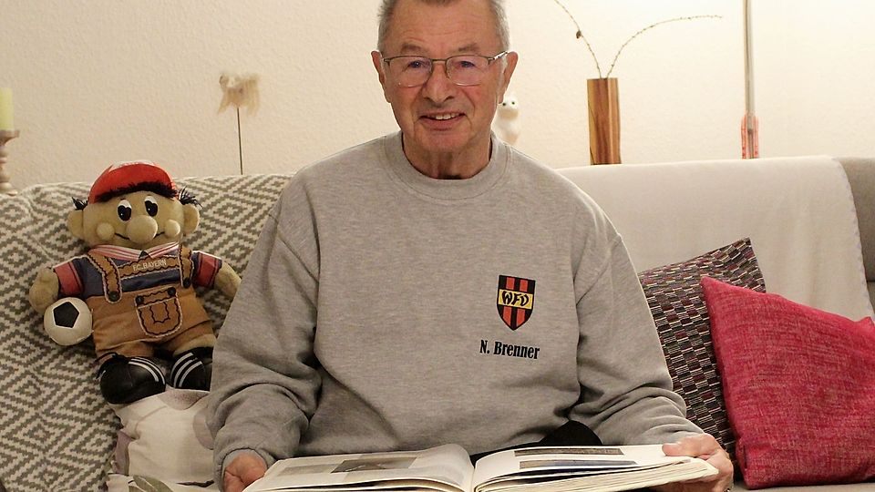 Norbert Brenner auf dem Sofa sitzend, in Erinnerungen blätternd und neben ihm Bayern Maskottchen "Bazi"