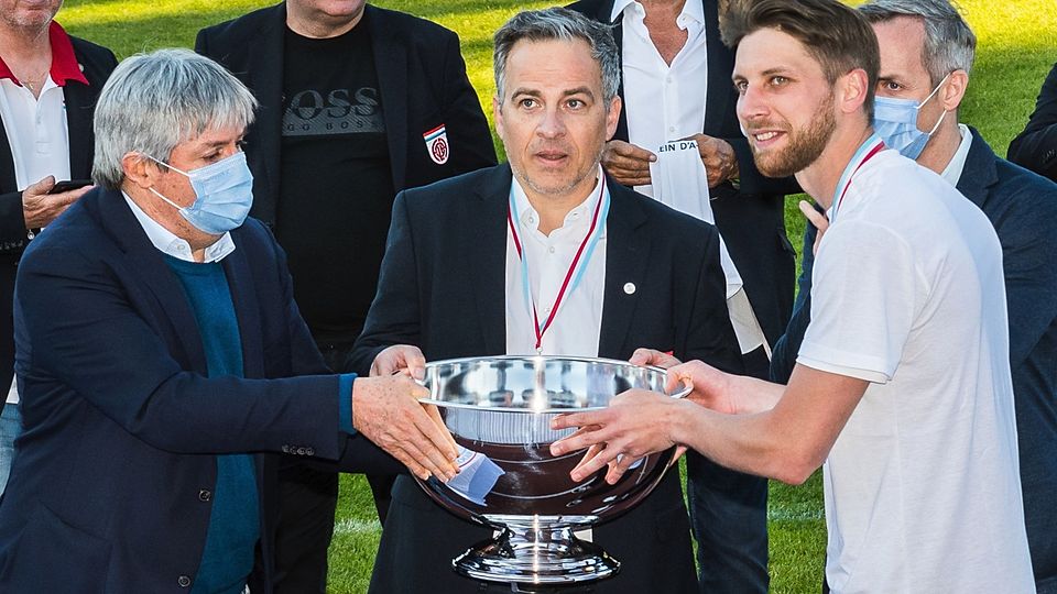Billy Bernard (r.) und Fola-Präsident Mauro Mariani (M.) bekommen den Meisterpokal vom FLF-Vorsitzenden Paul Philipp überreicht